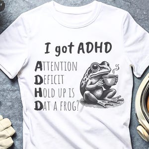 Pode incluir: Camiseta branca com texto preto que diz "Eu tenho TDAH" e um gr&aacute;fico de um sapo segurando uma x&iacute;cara de caf&eacute;. O texto abaixo do sapo diz "Attention Deficit Hold Up Is Dat A Frog?"
