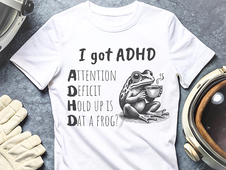 Pode incluir: Camiseta branca com texto preto que diz "Eu tenho TDAH" e um gr&aacute;fico de um sapo segurando uma x&iacute;cara de caf&eacute;. O texto abaixo do sapo diz "Attention Deficit Hold Up Is Dat A Frog?"