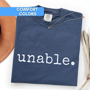 Op de afbeelding: Een marineblauw T-shirt met het woord "unable" in het wit gedrukt. Het shirt is opgevouwen en heeft een gouden ketting met een sterhanger. Een bruine hoed en gedroogde bladeren zijn ook in de afbeelding.