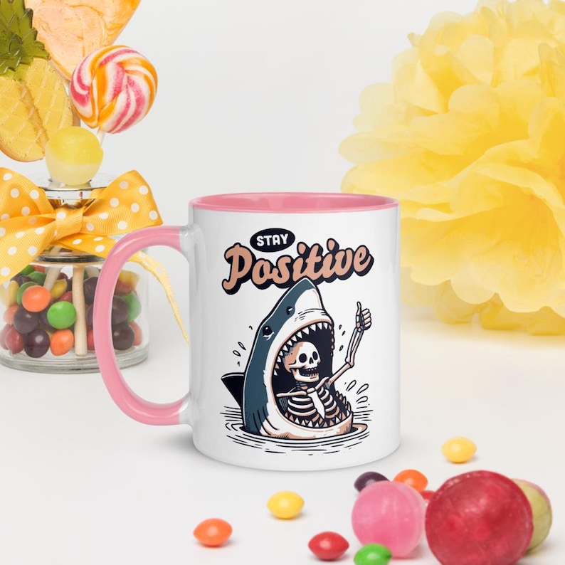 May include: White ceramic mug with a pink interior and handle. The mug features a cartoon shark design with a skeleton giving a thumbs up, and the words "Stay Positive". Candy and decorations are in the background.