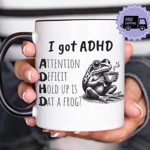 Pode incluir: Caneca de cerâmica branca com acabamento e alça pretos. A caneca apresenta o texto "I got ADHD" com um acrónimo e um desenho animado de um sapo segurando uma xícara de café. A caneca é segurada por uma pessoa.