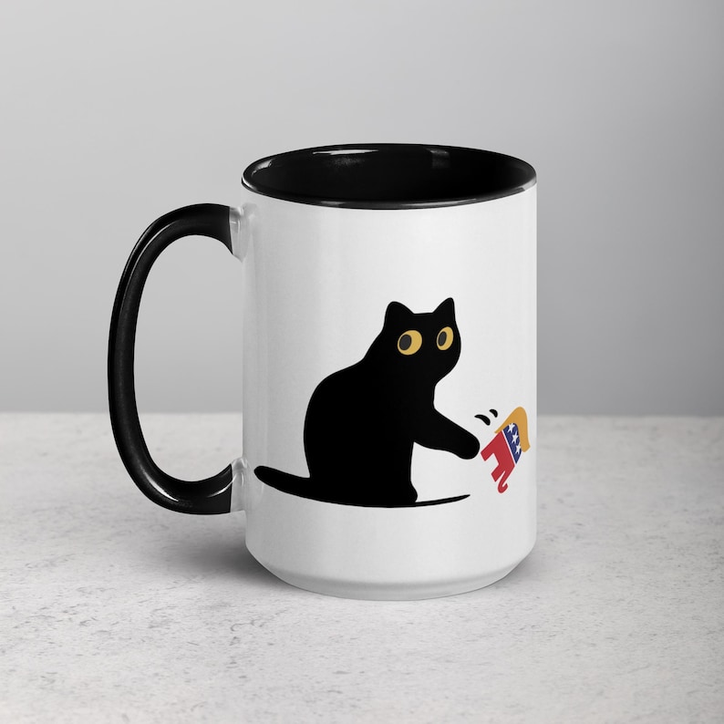 May include: A white ceramic mug with a black rim and handle. The mug features a black cat with yellow eyes swatting at a red, white, and blue elephant.