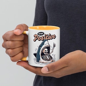 May include: White ceramic mug with a yellow interior and handle. The mug features a cartoon illustration of a shark with a skeleton inside, giving a thumbs up. The text "Stay Positive" is above the shark.