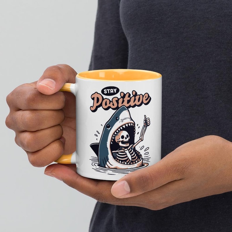 May include: White ceramic mug with a yellow interior and handle. The mug features a cartoon illustration of a shark with a skeleton inside, giving a thumbs up. The text "Stay Positive" is above the shark.