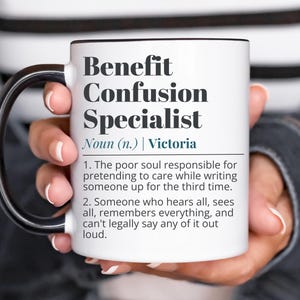 Op de afbeelding: Witte keramische mok met zwart handvat en rand. De mok toont de tekst "Benefit Confusion Specialist" en een definitie. De mok wordt door een persoon vastgehouden.