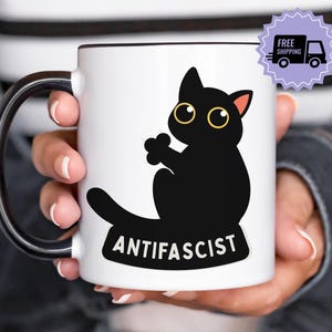 Könnte beinhalten: Eine weiße Keramiktasse mit schwarzem Rand und Henkel. Die Tasse zeigt eine schwarze Cartoon-Katzen-Illustration mit gelben Augen und rosa Ohren. Das Wort "ANTIFASCIST" ist darunter gedruckt.