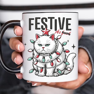Könnte beinhalten: Ein weißer Keramikbecher mit schwarzem Henkel und Rand, verziert mit einer Grafik einer mürrischen weißen Katze, die in rote und grüne Lichterketten gewickelt ist. Über der Katze steht der Text FESTIVE in fetten schwarzen Buchstaben.