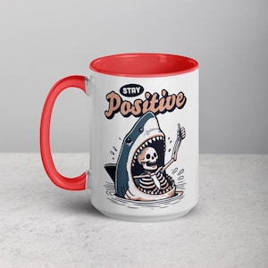 May include: White ceramic mug with a red interior and handle. The mug features a cartoon illustration of a shark with a skeleton inside, giving a thumbs-up. The text "STAY Positive" is above the shark.