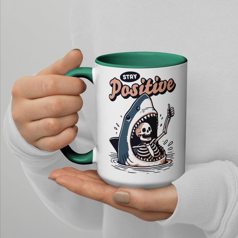 May include: White ceramic mug with a green handle and interior. The mug features a cartoon shark with a skeleton inside, giving a thumbs-up. The words "STAY Positive" are written above the shark in a retro font.