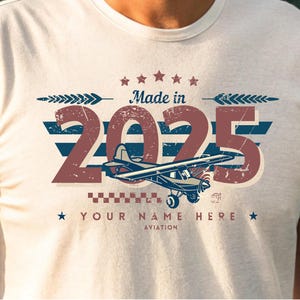 May include: White t-shirt with a vintage-style graphic design featuring a small airplane, the year "2025", and the text "Made in 2025". The design also includes stars, stripes, and a checkered pattern. The text "Your Name Here Aviation" is printed below the graphic.