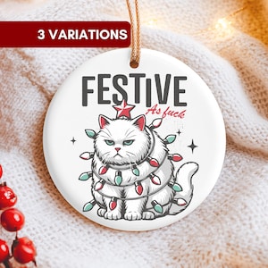 Könnte beinhalten: Weißer Keramik-Weihnachtsschmuck mit einem grummeligen Katzendesign. Der Schmuck zeigt den Text "FESTIVE As fuck" und eine Cartoon-Katze, die in Weihnachtsbeleuchtung eingewickelt ist. Ein goldener Faden ist zum Aufhängen befestigt.