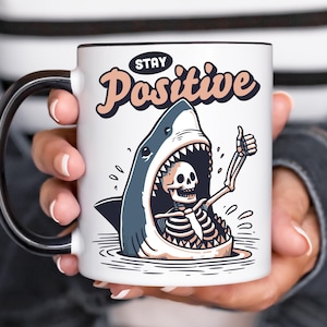 Puede incluir: Taza de cerámica blanca con asa y borde negros. La taza presenta una ilustración de dibujos animados de un tiburón con un esqueleto en su interior haciendo una señal de pulgar hacia arriba. El texto "Stay Positive" está encima del tiburón.