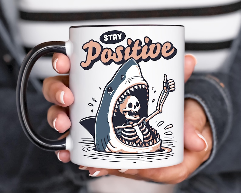 May include: White ceramic mug with a black handle and rim. The mug features a cartoon illustration of a shark with a skeleton inside giving a thumbs up. The text "Stay Positive" is above the shark.