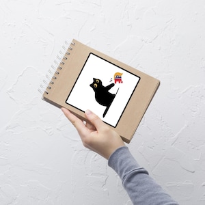 Puede incluir: Un cuaderno en espiral marr&oacute;n con una ilustraci&oacute;n de un gato negro sobre un cuadrado blanco. El gato se estira hacia un dibujo animado de un pol&iacute;tico con pelo rubio y un elefante rojo, blanco y azul.