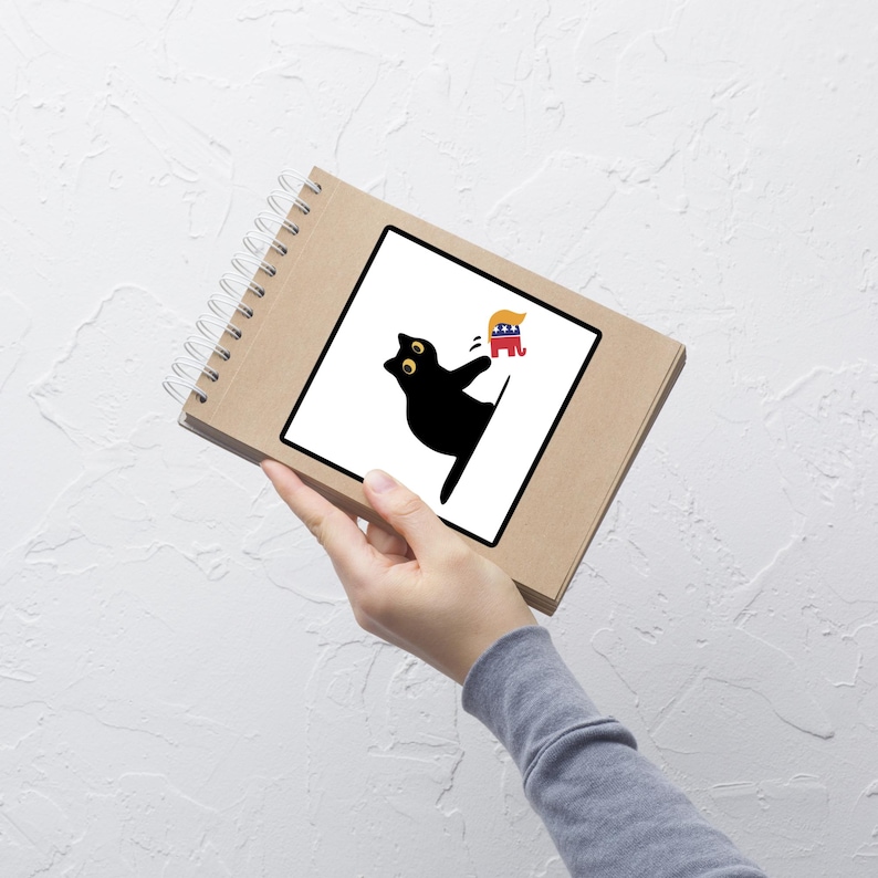 Puede incluir: Un cuaderno en espiral marr&oacute;n con una ilustraci&oacute;n de un gato negro sobre un cuadrado blanco. El gato se estira hacia un dibujo animado de un pol&iacute;tico con pelo rubio y un elefante rojo, blanco y azul.