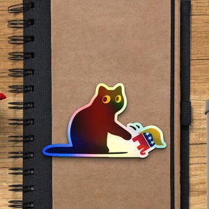 Puede incluir: Una pegatina hologr&aacute;fica de un gato golpeando a un elefante de dibujos animados con un dise&ntilde;o rojo, blanco y azul. La pegatina est&aacute; sobre un cuaderno marr&oacute;n con encuadernaci&oacute;n en espiral negra. L&aacute;pices y un recipiente met&aacute;lico en el fondo.