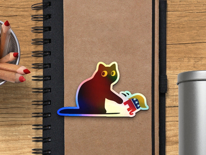 Puede incluir: Una pegatina hologr&aacute;fica de un gato golpeando a un elefante de dibujos animados con un dise&ntilde;o rojo, blanco y azul. La pegatina est&aacute; sobre un cuaderno marr&oacute;n con encuadernaci&oacute;n en espiral negra. L&aacute;pices y un recipiente met&aacute;lico en el fondo.
