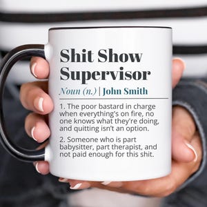 Op de afbeelding: Witte keramische mok met zwarte handgreep en rand, met de tekst "Shit Show Supervisor" en een humoristische definitie. De mok wordt door een persoon vastgehouden.