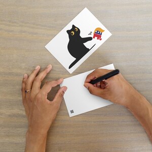 Puede incluir: Dos postales blancas sobre una superficie de madera. Una postal presenta una silueta de gato negro con ojos amarillos, golpeando a un elefante de dibujos animados con un tup&eacute; rubio. La otra postal est&aacute; en blanco, con una persona escribiendo en ella con un bol&iacute;grafo negro.