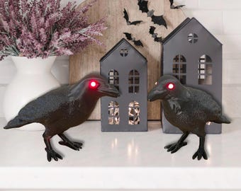 Puede incluir: Dos figuras de cuervos negros con ojos rojos brillantes. Detrás, dos candelabros grises con forma de casa y una tabla de cortar de madera con murciélagos recortados. Un jarrón blanco con flores moradas a la izquierda. Tema de Halloween.