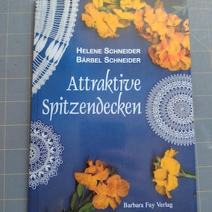 May include: Book cover with a blue background featuring white lace doilies and yellow flowers. The title of the book is "Attractive Spitzendecken" by Helene Schneider and Bärbel Schneider. The book is published by Barbara Fay Verlag.