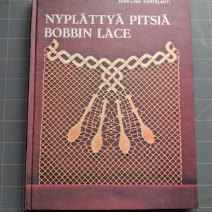 May include: A red book with the title "NYPLÄTTYÄ PITSIÄ BOBBIN LACE" and the author's name "EEVA-LIISA KORTELAHTI" printed on the cover. The cover features a detailed image of a lace pattern.