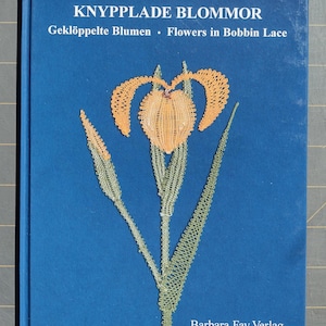 May include: A blue book with the title "KNYPPLADE BLOMMOR" in white text. The subtitle is "Geklöppelte Blumen - Flowers in Bobbin Lace". The book cover features a delicate orange and green bobbin lace flower.
