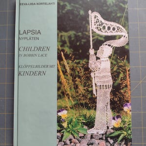 May include: A book titled "Lapsia Nyplaten Children in Bobbin Lace Kloppelbilder Mit Kindern" by Eeva-Liisa Kortelahti. The cover features a white bobbin lace figure of a child holding a flag.