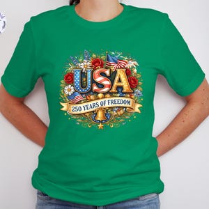 May include: Green t-shirt with a colorful graphic design. The design features the word "USA" in red, white, and blue, surrounded by flowers, flags, and the text "250 YEARS OF FREEDOM".
