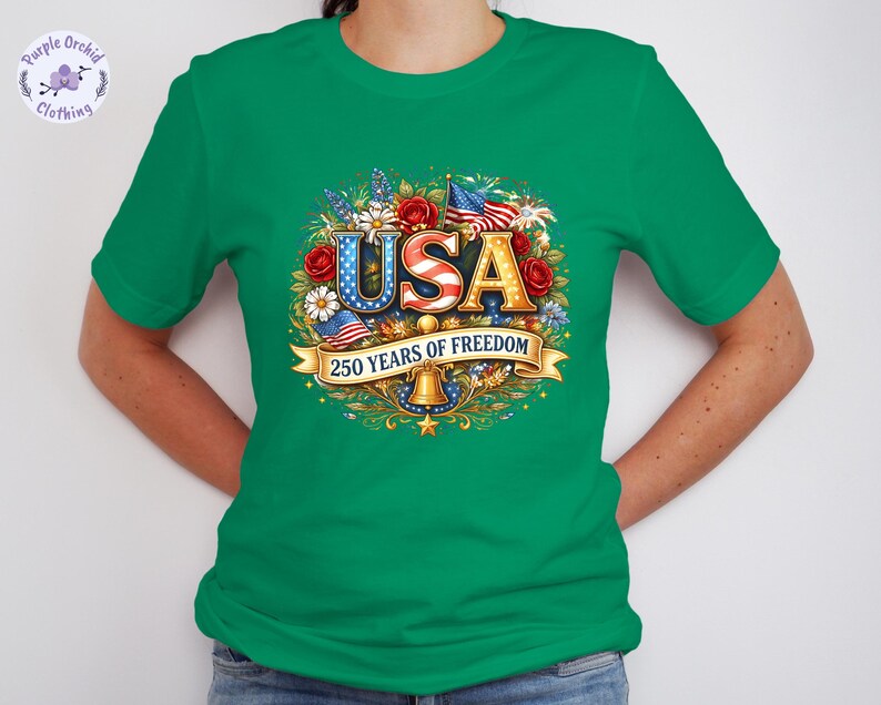 May include: Green t-shirt with a colorful graphic design. The design features the word "USA" in red, white, and blue, surrounded by flowers, flags, and the text "250 YEARS OF FREEDOM".