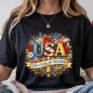 May include: Black t-shirt featuring a patriotic design with the word "USA" in red, white, and blue, surrounded by flowers, flags, and fireworks. The text "250 YEARS OF FREEDOM" is displayed on a banner below.
