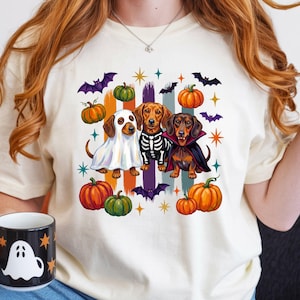 May include: A cream-colored t-shirt featuring a graphic of three dachshunds dressed in Halloween costumes: a ghost, a skeleton, and a vampire. The design includes orange and green pumpkins, purple bats, and gold stars against vertical brush strokes.