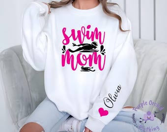 May include: White sweatshirt with the words "swim mom" in pink script and a black swimmer graphic. The sleeve features the name "Olivia" in script with a pink heart. Purple Orchid Clothing logo is visible.