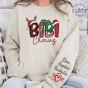 May include: Beige sweatshirt with the text "Bibi Claus" in a festive design. The letters are decorated with Christmas elements like a reindeer antler, a Christmas tree, and a Santa hat. The sleeve has the names Lisa, Sophia, and Isabella.