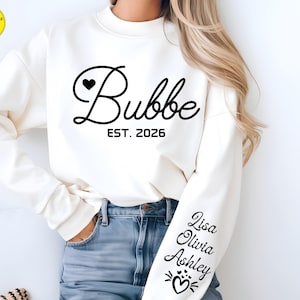 May include: Cream-colored sweatshirt with the word "Bubbe" in a cursive font, a heart, and the text "EST. 2026". The sleeve features the names "Lisa", "Olivia", and "Ashley" in a script font, along with heart and cat graphics.