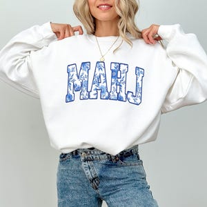 May include: White sweatshirt with the word "MAHJ" in blue floral print lettering. The sweatshirt is a crew neck style. The model is wearing blue jeans and a gold necklace.