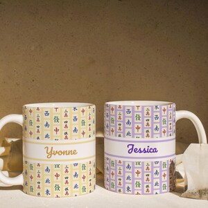May include: Two ceramic mugs with white handles. One mug is yellow with a white band and the name "Yvonne" in gold script. The other is lavender with "Jessica" in purple script. Both mugs have a repeating pattern of Chinese characters.