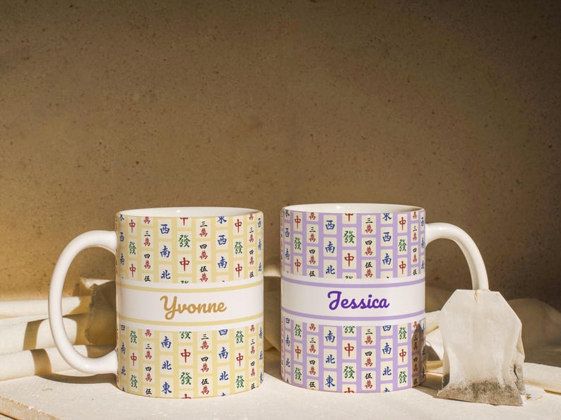 May include: Two ceramic mugs with white handles. One mug is yellow with a white band and the name "Yvonne" in gold script. The other is lavender with "Jessica" in purple script. Both mugs have a repeating pattern of Chinese characters.