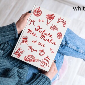 May include: A white notebook with red illustrations and text, including a disco ball, flowers, doves, and a ring. The cover reads "Future Mrs. Martin est. 2026" in red script. The notebook is held on a person's lap.