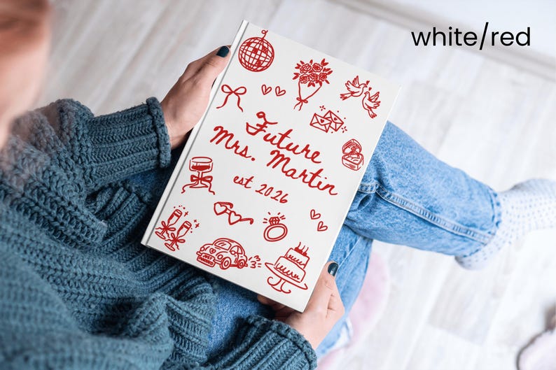 May include: A white notebook with red illustrations and text, including a disco ball, flowers, doves, and a ring. The cover reads "Future Mrs. Martin est. 2026" in red script. The notebook is held on a person's lap.