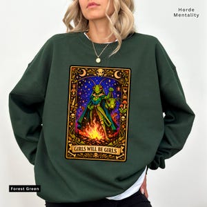 May include: Forest green sweatshirt with a graphic featuring a grasshopper-like figure in a cape holding a lantern over a fire, with the text "GIRLS WILL BE GIRLS". The design is framed with celestial and skull motifs.