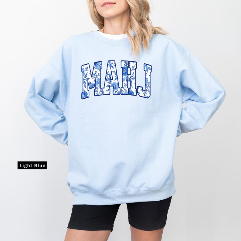 May include: Light blue sweatshirt with the word "MAHJ" in a blue floral design. The sweatshirt is a light blue color and has a crew neck. The text "Light Blue" is in a black rectangle in the lower left corner.