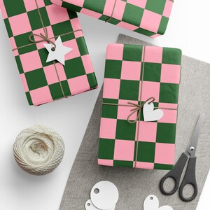 May include: Overhead view of wrapped presents with pink and green chequered paper. Each gift is tied with twine and has a white tag in the shape of a star or heart. A ball of twine, scissors, and blank tags are also visible.