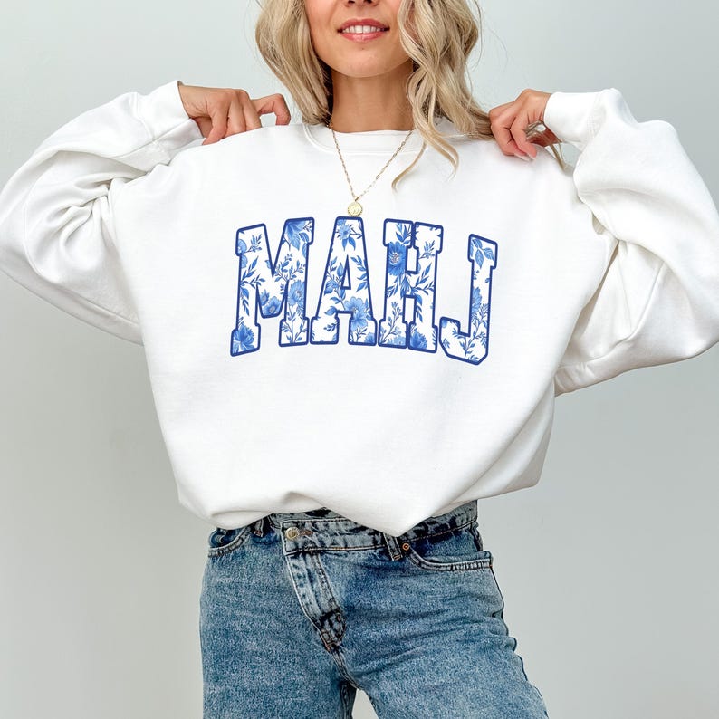 May include: White sweatshirt with the word "MAHJ" in blue floral print lettering. The sweatshirt is a crew neck style. The model is wearing blue jeans and a gold necklace.