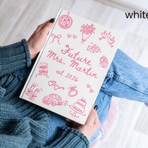 May include: A white notebook with pink illustrations and text. The cover reads "Future Mrs. Martin est. 2026" with drawings of a disco ball, flowers, and a cake. The notebook is held by a person wearing a blue sweater and jeans.