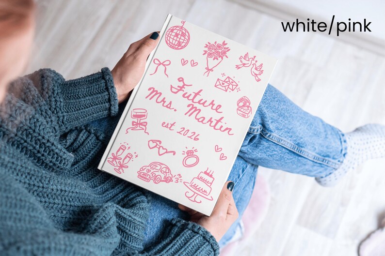 May include: A white notebook with pink illustrations and text. The cover reads "Future Mrs. Martin est. 2026" with drawings of a disco ball, flowers, and a cake. The notebook is held by a person wearing a blue sweater and jeans.