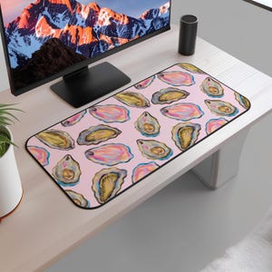 May include: A rectangular mouse pad featuring a pattern of watercolor oyster illustrations in shades of pink, yellow, and blue on a light pink background. The mouse pad has a black border and is placed on a white desk.