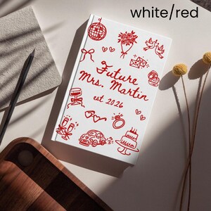May include: A white notebook with red illustrations and text that reads "Future Mrs. Martin est. 2026." The notebook is decorated with drawings of wedding-related items, including a cake, ring, and champagne glasses.