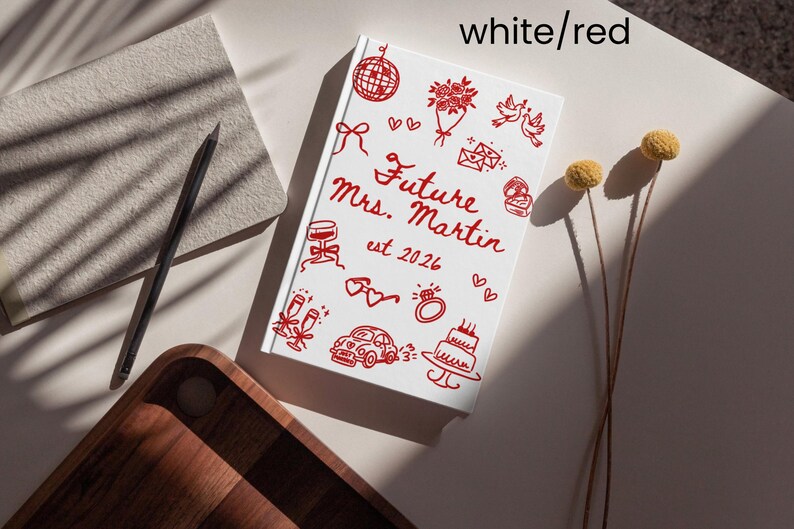 May include: A white notebook with red illustrations and text that reads "Future Mrs. Martin est. 2026." The notebook is decorated with drawings of wedding-related items, including a cake, ring, and champagne glasses.
