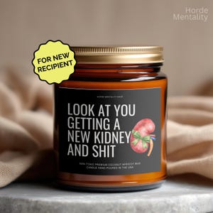 May include: A candle in a glass jar with a gold lid. The label reads "LOOK AT YOU GETTING A NEW KIDNEY AND SHIT" with a kidney graphic. A yellow sticker says "FOR NEW RECIPIENT". The candle is hand-poured in the USA.